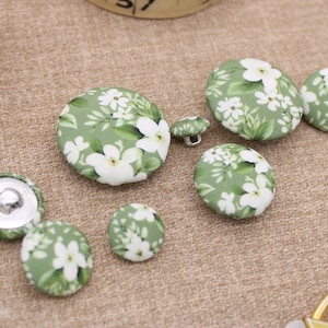 May include: A selection of fabric-covered buttons in different sizes. The buttons have a green base with a white floral design. A reel of red thread and scissors are also in the image.