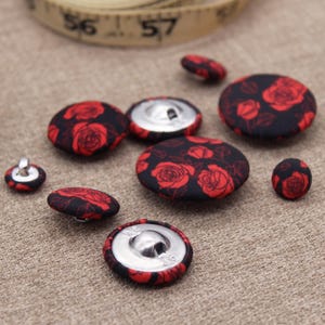 May include: Various fabric-covered buttons featuring a red rose pattern on a black background. The buttons have silver metal backs and come in different sizes. A blue thread spool and a measuring tape are in the background.