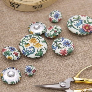 May include: Various fabric-covered buttons with floral designs in red, blue, and yellow on a cream base. The buttons are of different sizes and are arranged on a beige surface, with a thread spool and scissors nearby.