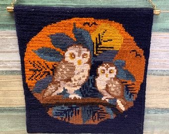 Op de afbeelding: Een blauwe wandkleed met twee bruine uilen die op een tak zitten. De uilen zijn omringd door oranje en gele bladeren en een volle maan. Het wandkleed hangt aan een gouden ketting.