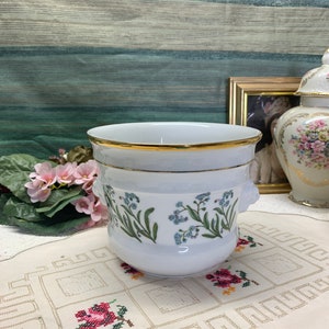 Op de afbeelding: Witte keramische plantenpot met een gouden rand en een bloemenontwerp met blauwe vergeet-me-nietjes. De pot heeft twee kleine leeuwenkopgrepen aan de zijkanten. De pot staat op een wit gehaakt tafellaken.