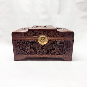 May include: A rectangular, dark brown wooden box with intricate carvings. The box features a gold-colored clasp and detailed relief carvings of figures and foliage. The top of the box has a decorative carved design. The box is set against a white background.