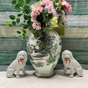 Puede incluir: Dos figuritas de caniche de porcelana blanca que sostienen flores rosas, flanqueando un jarrón de cerámica blanca con un patrón floral verde y amarillo.