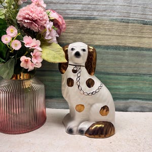 May include: A white ceramic dog figurine featuring metallic gold-toned ears, spots, and a painted chain collar. The dog is depicted in a seated position with a smooth, glossy finish, showcasing detailed facial features and a distinct metallic lustre.