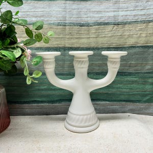 May include: White ceramic candle holder with three arms. The holder has a twisted rope design around the base.