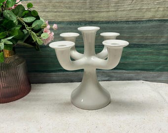 May include: A white ceramic candelabra with five candle holders. The candelabra has a unique, modern design with a wide base and five arms that extend out from the center.