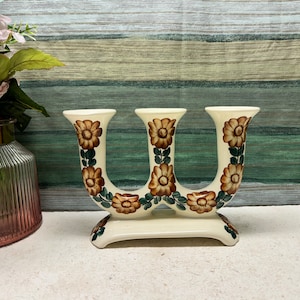 Peut inclure: Un bougeoir en céramique blanche avec trois coupelles pour bougies. Le bougeoir est décoré de motifs floraux marrons et verts.