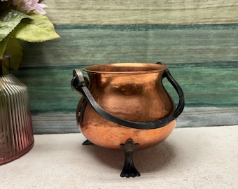 Puede incluir: Una pequeña olla de cobre con un acabado martillado y asas y patas de hierro negro. La olla está sobre una superficie de color marrón claro.