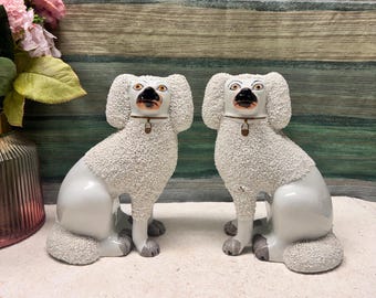 May include: A pair of white ceramic poodle figurines featuring textured, curly fur detailing on the head, chest, and tail. Each dog has a smooth body, painted facial features, and a gold-colored collar with a small hanging charm around the neck.