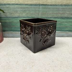 Könnte beinhalten: Ein dunkelbrauner, quadratischer Keramik-Pflanztopf mit geprägten Blumendesigns. Der Pflanztopf hat eine glänzende Oberfläche und eine rechteckige Form. Das Blumenmuster wiederholt sich auf mehreren Seiten.