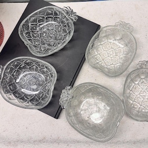 Pode incluir: Cinco tigelas transparentes em forma de abacaxi. As tigelas têm uma superfície texturizada e um topo de abacaxi detalhado. As tigelas estão dispostas sobre uma superfície preta, com um vaso rosa ao fundo. Estas tigelas são perfeitas para servir lanches ou sobremesas.