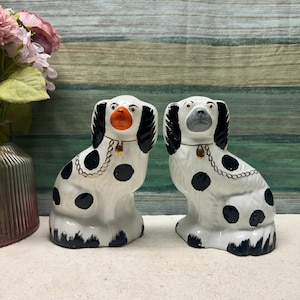 Puede incluir: Dos figuras de perro de cerámica. Los perros son blancos con manchas y detalles en negro, incluyendo orejas y patas negras. Cada perro tiene un collar de cadena dorada y una pequeña nariz de color naranja. Las figuras se exhiben sobre una superficie clara.
