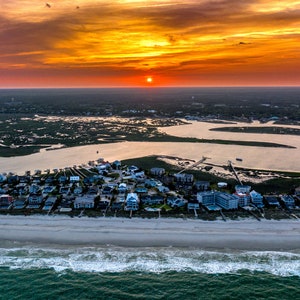 May include: Aerial view of a coastal town with houses lining a sandy beach. The ocean is in the foreground with waves crashing on the shore. A river winds through the town, leading to a marsh in the background. The sky is a vibrant orange and red, indicating a sunset.