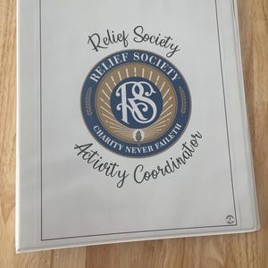 Puede incluir: Una carpeta blanca con un logotipo dorado y azul que dice "Relief Society" y "Charity Never Faileth". La carpeta también dice "Activity Coordinator".