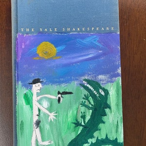 May include: A blue book with the text "THE YALE SHAKESPEARE" on the cover. The cover features a hand-painted illustration of a person in a hat holding a gun, facing a green monster in a field of green grass.
