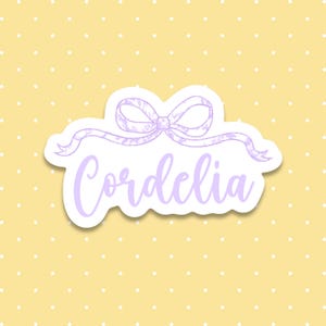 May include: A die-cut sticker featuring the name Cordelia written in a light purple cursive script font. Above the text is a light purple hand-drawn style ribbon bow with a subtle floral pattern. The sticker has a white border and shadow.