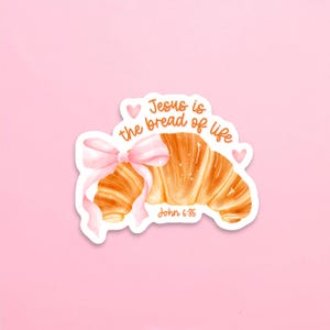May include: A sticker featuring a watercolor illustration of a croissant tied with a pink bow. The text "Jesus is the bread of life" and "John 6:35" are written in orange script, with pink hearts. The background is a solid light pink.