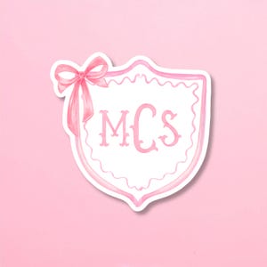 May include: A pink and white sticker featuring a shield design with the letters "MCs" in a decorative font. A pink bow is at the top left. The sticker has a scalloped border and is set against a pink background.