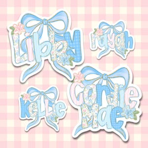 May include: Four decorative stickers featuring the names Libby, Rayah, Kellie, and Gordie Mae. Each name is written in a stylized font with blue, pink, and floral patterns, accented by a large light blue bow and small pink and blue flowers.