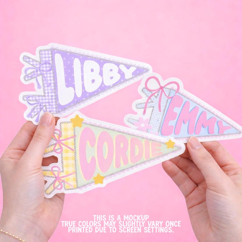Pode incluir: Tr&ecirc;s autocolantes triangulares em forma de galhardete com os nomes LIBBY, EMMY e CORDIE em letras a negrito. Os designs incluem padr&otilde;es axadrezados, la&ccedil;os e estrelas em tons pastel roxo, azul e amarelo sobre um fundo rosa claro.