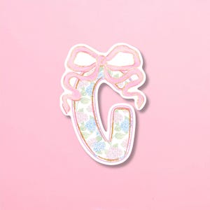 May include: A sticker shaped like the capital letter G, featuring a floral pattern of blue and pink hydrangea blossoms. The letter is outlined in a gold glitter effect and topped with a large, light pink ribbon bow with gold glitter edges.