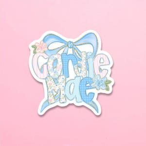May include: A die-cut sticker featuring the text "Gardie Mae" in a stylized font. The letters are filled with light blue and pink patterns, including stripes, gingham, and floral prints. A large light blue bow sits behind the text.