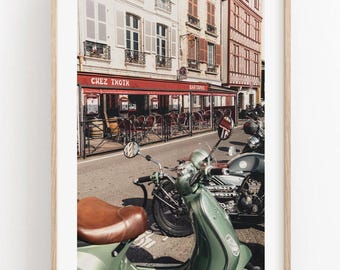 Op de afbeelding: Een ingelijste kunstdruk met een straatscène met een lichtgroene scooter op de voorgrond, een donkere motorfiets erachter en een restaurant met een rode luifel met het opschrift Chez Txotx, tegenover meerverdiepingsgebouwen.