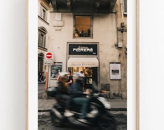 May include: A framed wall art print depicting a street scene in front of a Gelateria Florerè shop with a cream-colored awning. A blurred dark scooter with two riders moves across the foreground, while a window above shows an interior view.