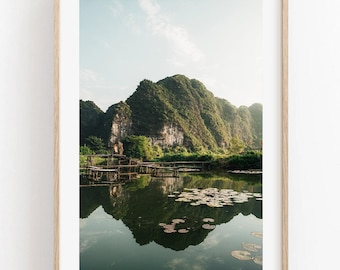 May include: A framed landscape print featuring a large, craggy green mountain reflected in a calm body of water. The foreground shows a wooden structure extending into the water, which is dotted with numerous lily pads under a bright sky.