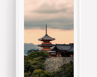 May include: A multi-tiered orange and dark gray pagoda stands atop a stone wall surrounded by lush green trees under a soft sunset sky. The structure features traditional curved rooflines and intricate architectural details, set against distant mountains.
