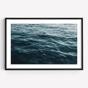 Può includere: Un'opera d'arte da parete incorniciata che presenta una fotografia dall'alto di acqua oceanica blu profondo con onde dolci e ondulate. L'immagine è racchiusa in una semplice cornice rettangolare nera sottile con un ampio bordo bianco.