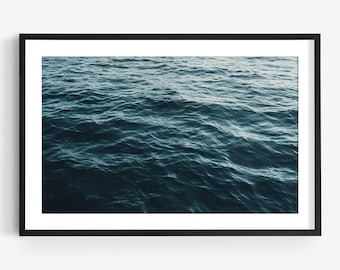 May include: A framed wall art piece featuring a high-angle photograph of deep blue ocean water with gentle, rolling waves. The image is encased in a simple, thin black rectangular frame with a wide white mat border surrounding the water print.
