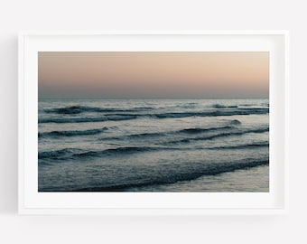 May include: A framed landscape photograph of the ocean at twilight. The scene features dark blue waves rolling toward the shore under a soft, gradient sky that transitions from a pale peach hue at the horizon to a muted gray tone above.