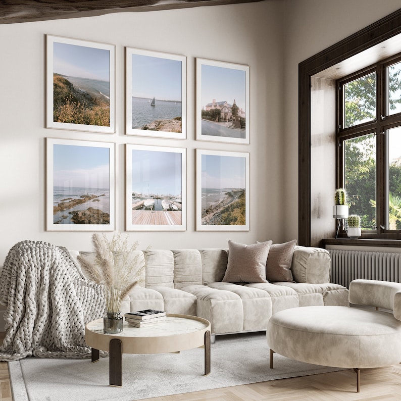 May include: A living room with a white couch, a round coffee table, and a white ottoman. There are six framed pictures on the wall, all featuring coastal scenes. The room has a large window with a view of trees outside.