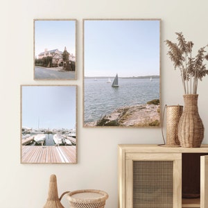May include: Three framed photographs of coastal scenes. The top left photo shows a building with a white facade and a blue roof. The top right photo shows a sailboat on a body of water with a rocky shore in the foreground. The bottom photo shows a wooden dock with several boats moored at the end.