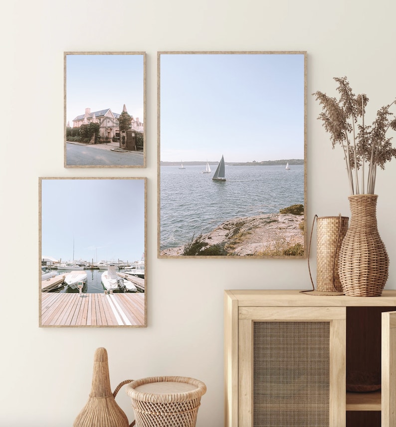 May include: Three framed photographs of coastal scenes. The top left photo shows a building with a white facade and a blue roof. The top right photo shows a sailboat on a body of water with a rocky shore in the foreground. The bottom photo shows a wooden dock with several boats moored at the end.