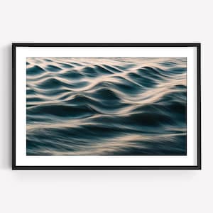 Puede incluir: Una impresión enmarcada que presenta una vista de primer plano de las olas del océano. El agua es de un azul profundo con reflejos blancos y dorados, creando una sensación de movimiento y textura. La obra de arte está enmarcada en un marco negro.
