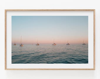 May include: A framed landscape photograph depicting several sailboats floating on calm, dark blue water under a soft, gradient sky that transitions from a pale blue at the top to a warm, light pink hue near the horizon line at sunset.