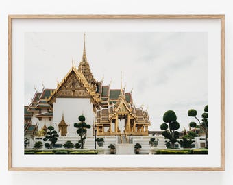 May include: A framed wall art print featuring a white temple building with intricate gold roof details and tiered spires. The structure is surrounded by manicured topiary trees and set against a pale, overcast sky in a light wood frame.