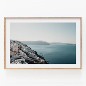 May include: A framed landscape photograph featuring a coastal town with white buildings perched on a dark cliffside overlooking a deep blue sea. Small sailboats are visible on the water under a clear, pale sky, all encased in a light wood frame.