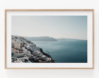 May include: A framed landscape photograph featuring a coastal town with white buildings perched on a dark cliffside overlooking a deep blue sea. Small sailboats are visible on the water under a clear, pale sky, all encased in a light wood frame.