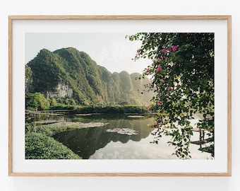 May include: A framed landscape photograph featuring a calm lake reflecting large, green, craggy mountains under a soft sky. In the foreground, a leafy branch with bright pink flowers hangs over the water, with green vegetation along the shoreline.