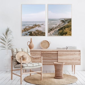 May include: Two framed photographs of a coastal scene with a rocky shoreline and ocean waves. The photos are hanging on a white wall above a wooden dresser with a woven rug on the floor.