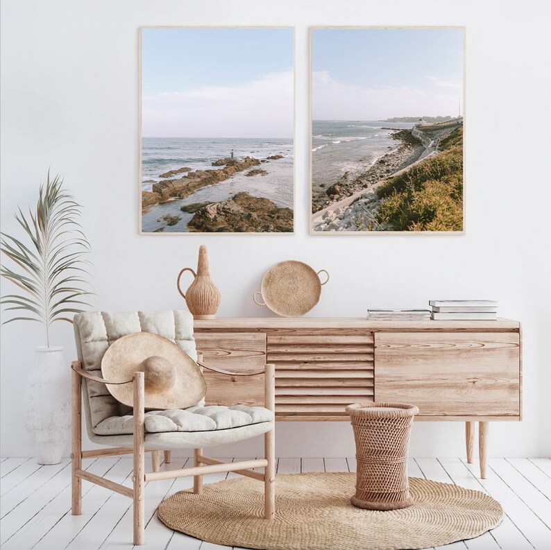 May include: Two framed photographs of a coastal scene with a rocky shoreline and ocean waves. The photos are hanging on a white wall above a wooden dresser with a woven rug on the floor.