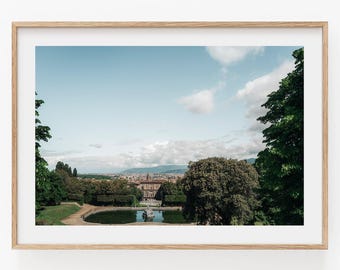 May include: A framed landscape photograph featuring a view of a historic building with a central fountain and pond, surrounded by lush green trees under a clear blue sky with scattered white clouds, all encased in a light-colored wooden frame.