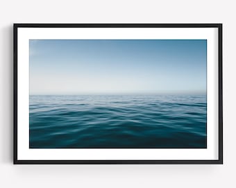 May include: A framed wall art print featuring a vast ocean scene with deep blue water transitioning into a lighter blue horizon under a clear sky. The image is enclosed in a simple black rectangular frame with a white mat border surrounding the print.