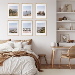 May include: Six framed photographs of coastal scenes, including a sailboat on the water, a house on a hill, and a rocky beach. The photos are hung on a white wall in a bedroom setting with a bed, a desk, and a chair.