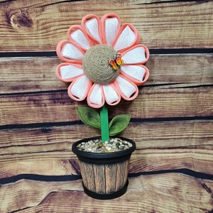May include: A pink and white fabric flower with a burlap center and two butterflies is in a black and brown planter with small stones. The planter has a wood grain design.
