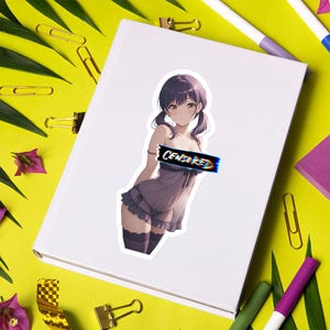 Può includere: Un adesivo con un'illustrazione in stile anime di una persona con i capelli viola in un abito viola chiaro. Il testo "CENSORED" è sul petto. L'adesivo è su una superficie bianca, circondato da forniture per ufficio su sfondo giallo.