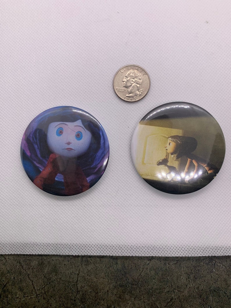 K&ouml;nnte beinhalten: Zwei runde Anstecknadeln mit Figuren aus dem Animationsfilm Coraline. Die erste Nadel zeigt Coraline mit ihren Knopfaugen, und die zweite Nadel zeigt Coraline, die in einem Raum mit einer T&uuml;r steht.